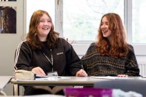 Two students sat side-by-side at a classroom table are laughing and smiling.