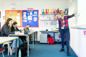 A maths tutor stands at the front of the class pointing towards the whiteboard. The class write in their books.