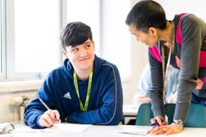 A maths tutor leans on a classroom table to help a student with their work.