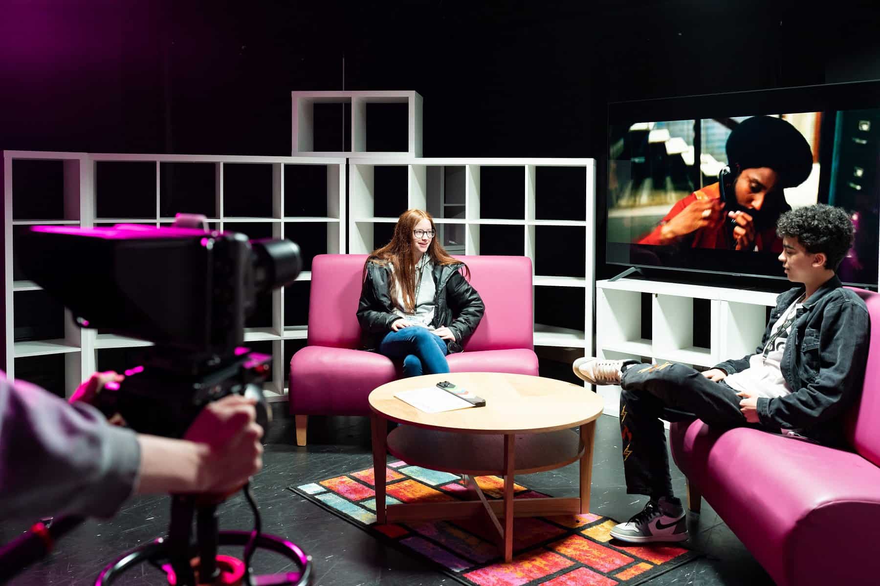 Two students sit in a TV studio with a large TV screen behind them. A camera is focused on them in the foreground.