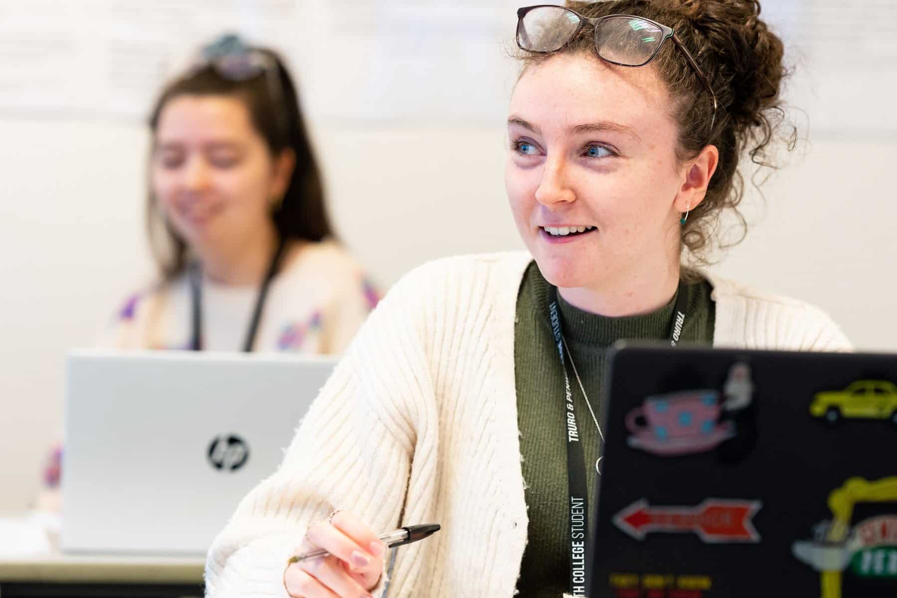 The camera focuses on a student who has their hair tied up, a pair of glasses on their head, a cream cardigan and a laptop open in front of them. They smile at someone we can't see.