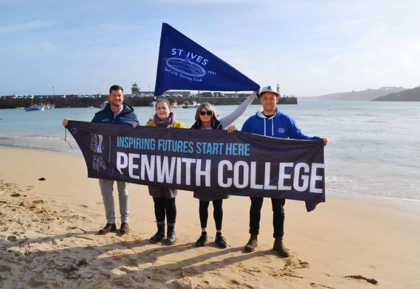 Members of staff from Penwith College stand on the beach at St Ives holding two banners which say St Ives surf life saving club. The other sign says inspiring futures start here.