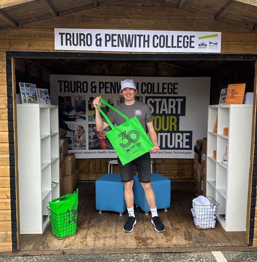 A Truro and Penwith College staff member at the stand at Boardmasters festival. He is holding a green tote bag and has other literature around him.