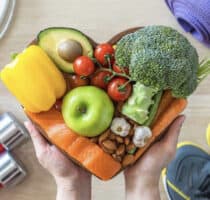 A pair of hands hold a heart-shaped, wooden chopping board with an assortment of food products on it.