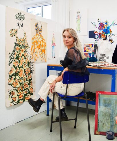 Young woman sits in her studio surrounded by colourful artwork.