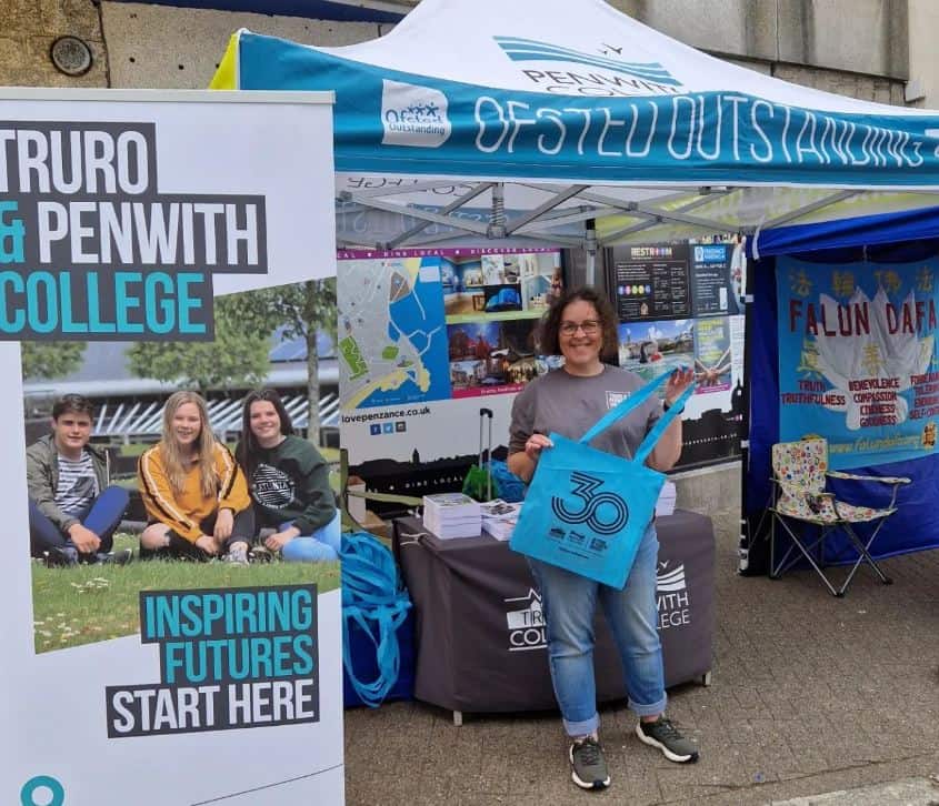 A member of staff from Penwith College stands at their table at the Mazey day celebrations in Penzance. They are holding a goodie bad and have a pull up next to them.