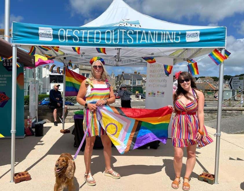 Two members of staff from Penwith College hold a rainbow flag in front of their stall at Penzance Pride 2023. They are dressed in rainbow wear and have a dog with them on a lead.