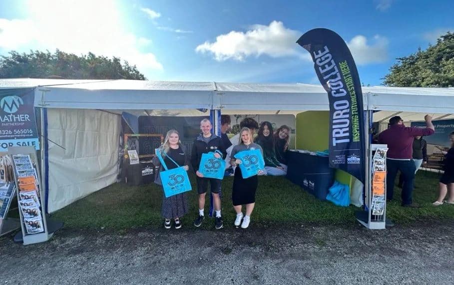 Three members of staff from Truro and Penwith College stand outside their stand at Stithian's show 2023. They are holding up blue, branded goodie bags.