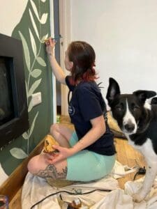 a girl is sitting in front of a green wall painting white leaves. There is a black and white dog behind her.