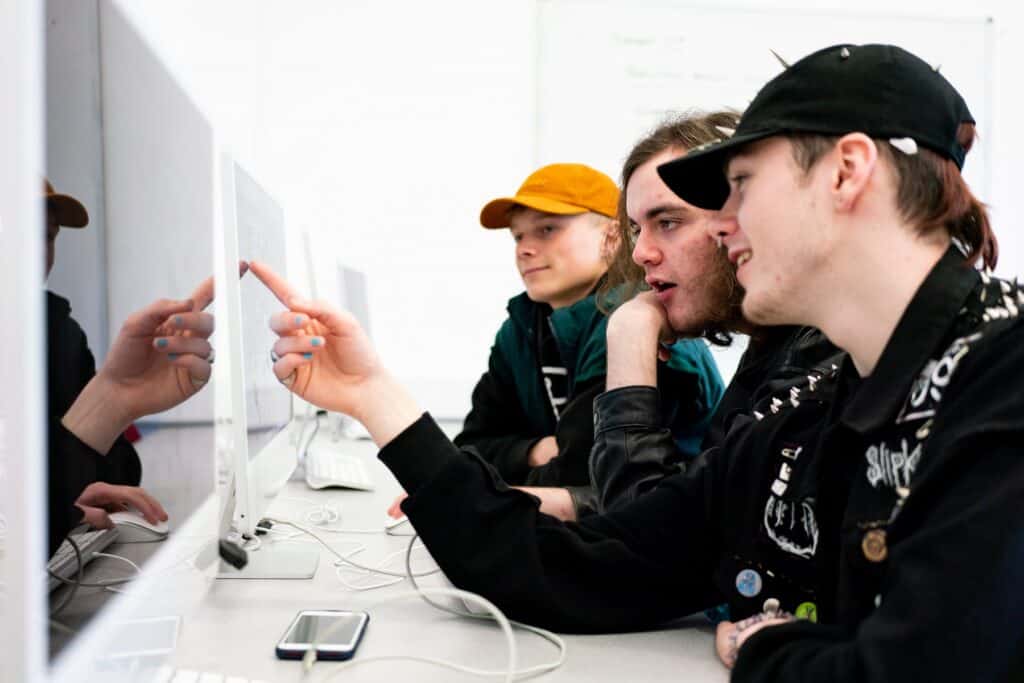 Three students sit at apple computers looking at the screens together.