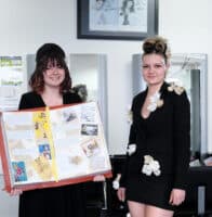 Two hairdressing students smile holding their mood boards and show their final designs.