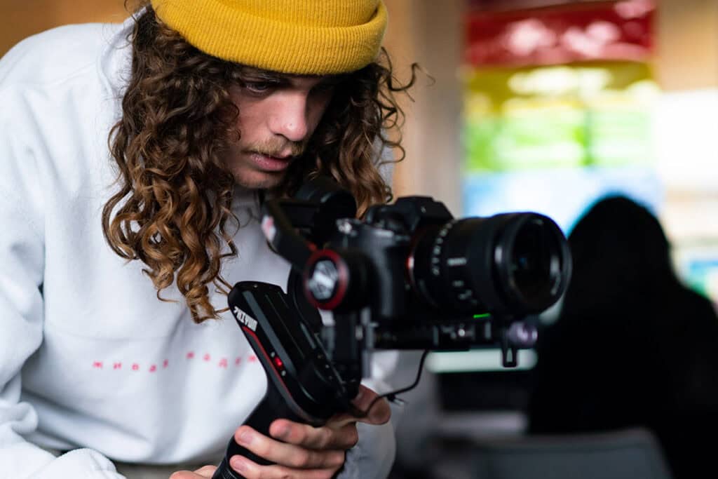 A student with a white jumper and yellow beanie hat looks closely through a digital camera.