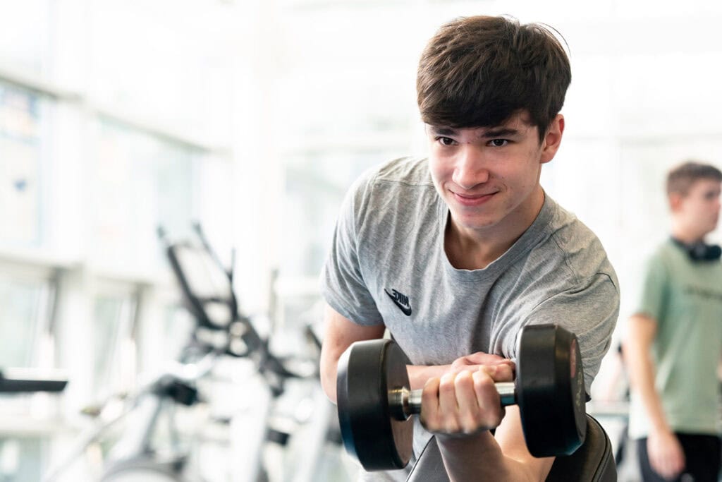 An image of a student at the college's gym. He is wearing a grey shirt and lifting a small weight while resting his arm.