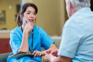 Nurse attentively listens to senior patient in nursing home. She is holding a clipboard in her lap and wearing scrubs. The man's back is to the camera.