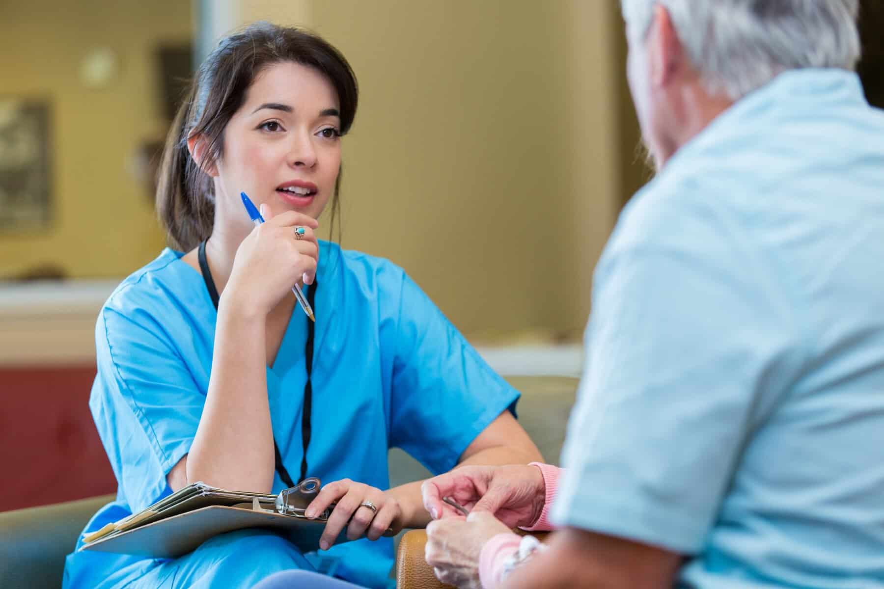 Nurse attentively listens to senior patient in nursing home. She is holding a clipboard in her lap and wearing scrubs. The man's back is to the camera.