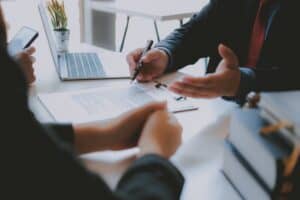 a close up of hands belong to three people sat around a desk. There is also a clipboard and pen and laptop on the desk.