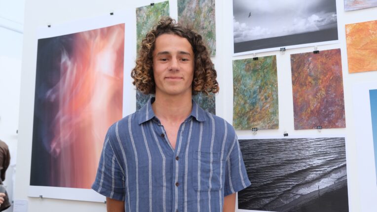 Young male student wearing a blue shirt and has curly brown hair is standing in front of his landscape paintings.