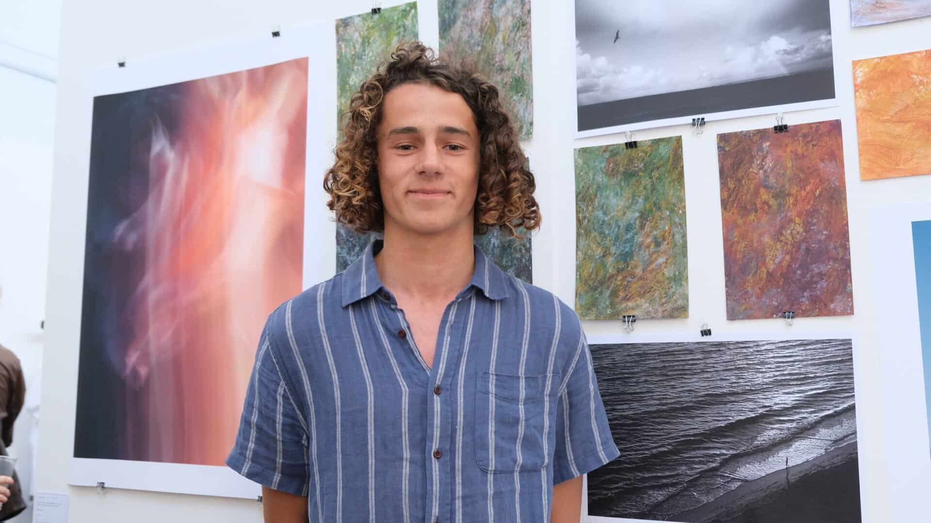 Young male student wearing a blue shirt and has curly brown hair is standing in front of his landscape paintings.