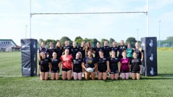 The female rugby academy in a group photo on a rugby pitch underneath the goal posts.