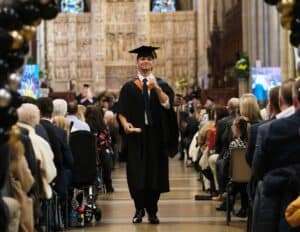 A male Graduate fist pumps the air while walking down the centre aisle of the Cathedral with his scoll while wearing cap and gown