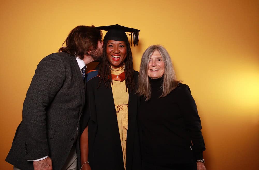 Grad 24 Photobooth A graduate in cap and gown receives a kiss on the cheek from one of her guests on her right while another smiles down camera to her left infront of the gold backdrop.