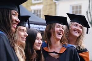 A group of students in Graduation robes smile and laugh towards an unknown person while posing for a photo.