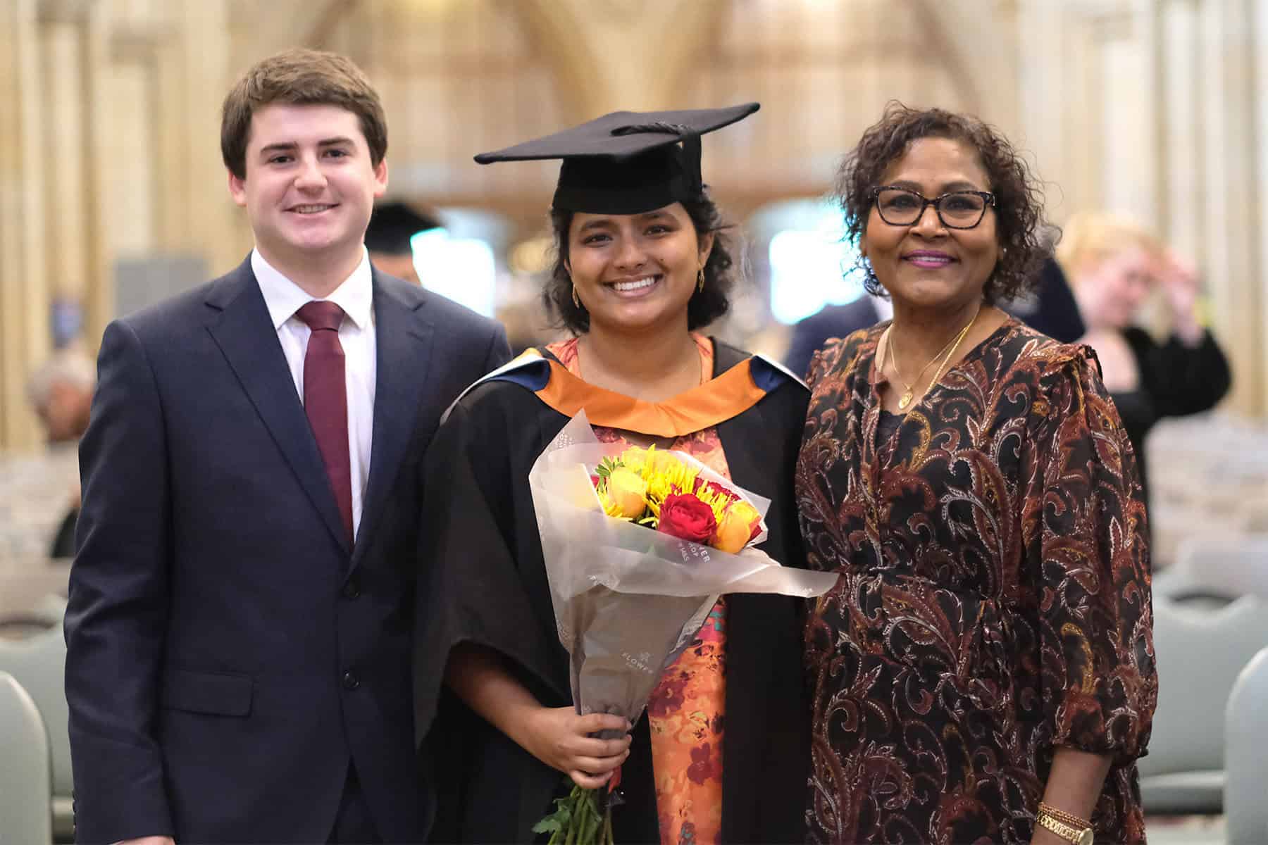 Graduation 24 05 A student in robes holds a bunch of flowers and poses for a photo with two guests inside Truro Cathedral.