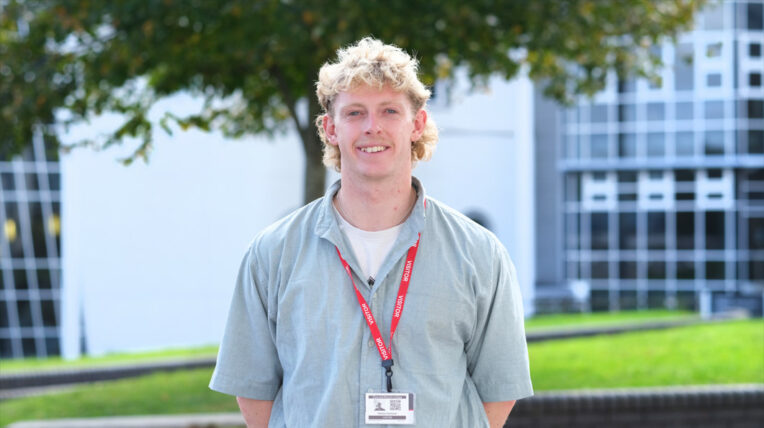 Man, wearing a shirt with short curly blonde hair smiles at the camera. In the background is the college campus.