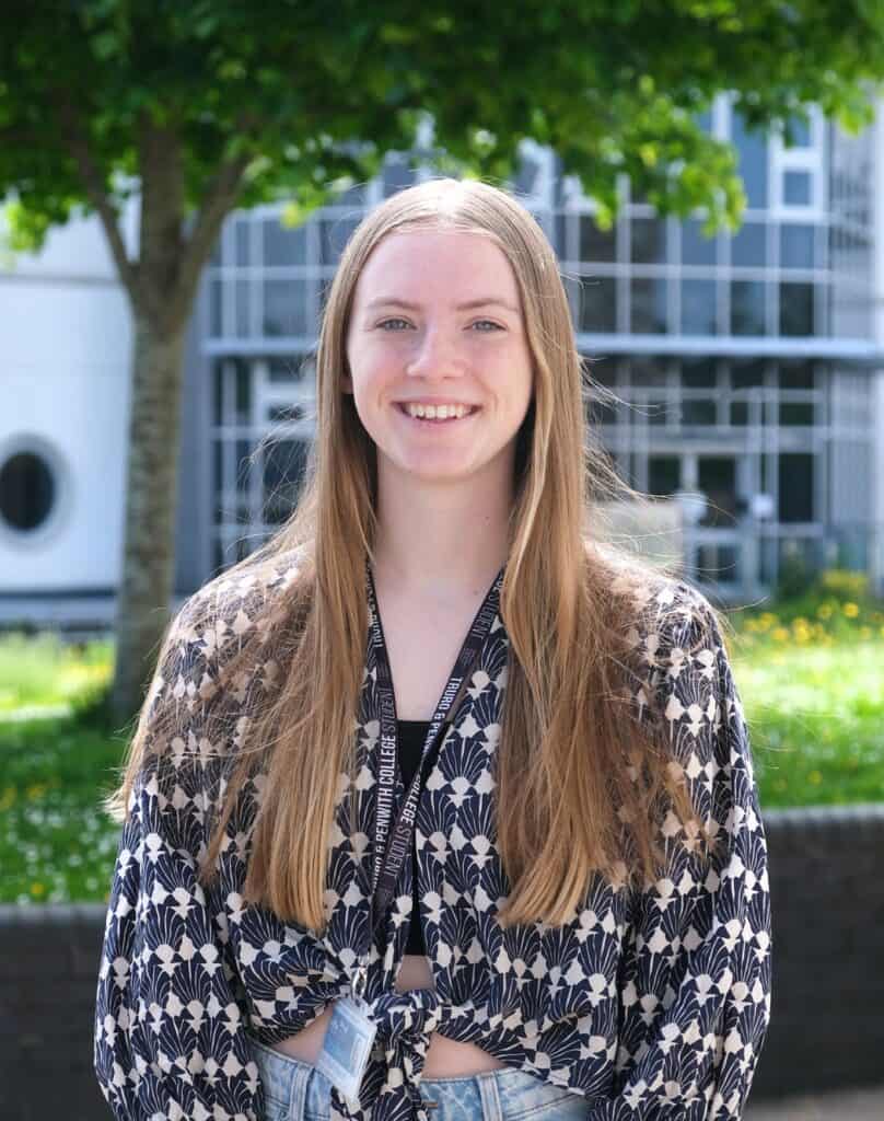 Young woman with long llight brown hair is wearing a checkered long sleeve top and is smiling at the camera with college campus in the background.