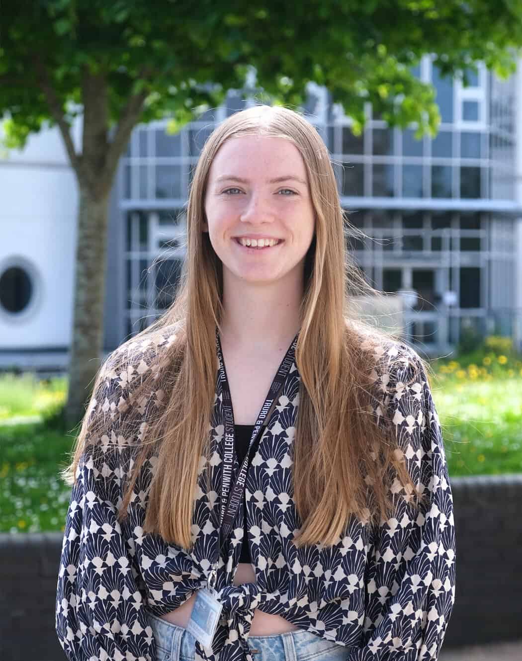 Young woman with long llight brown hair is wearing a checkered long sleeve top and is smiling at the camera with college campus in the background.