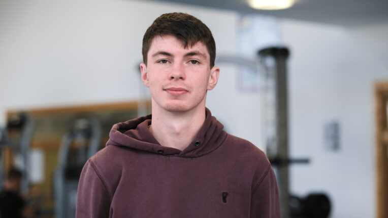Young man with a burgundy hoodie on and short brown hair is looking at the camera.