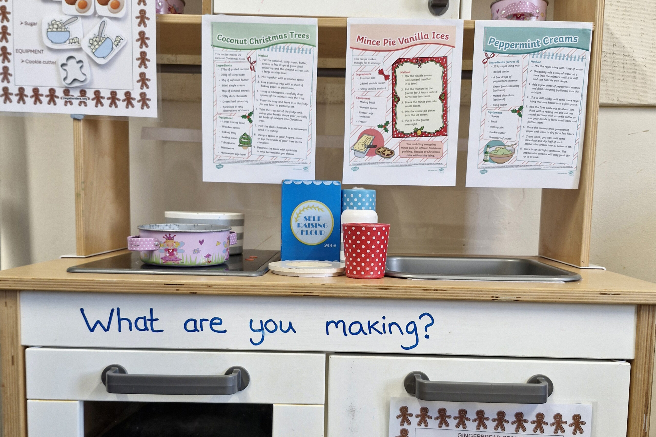 A toy kitchen has the words 'what are you making?' on it. There are three christmas recipes stuck to the top shelf.