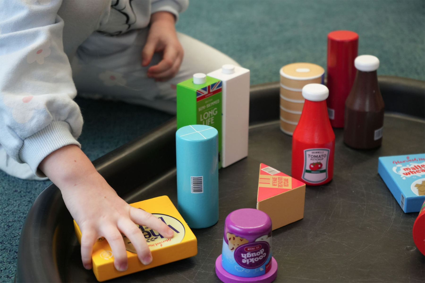A collection of wooden toys that mimic food cupboard items are layed out on a tray. A child sat next to them reaches out to one of the toys.