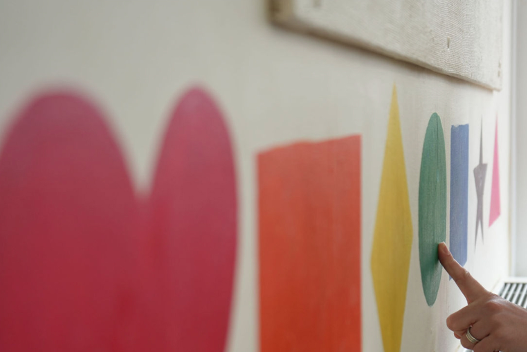 A close-up of a hand pointing to a green circle painted on the wall. There are other shapes of different colours alongside.