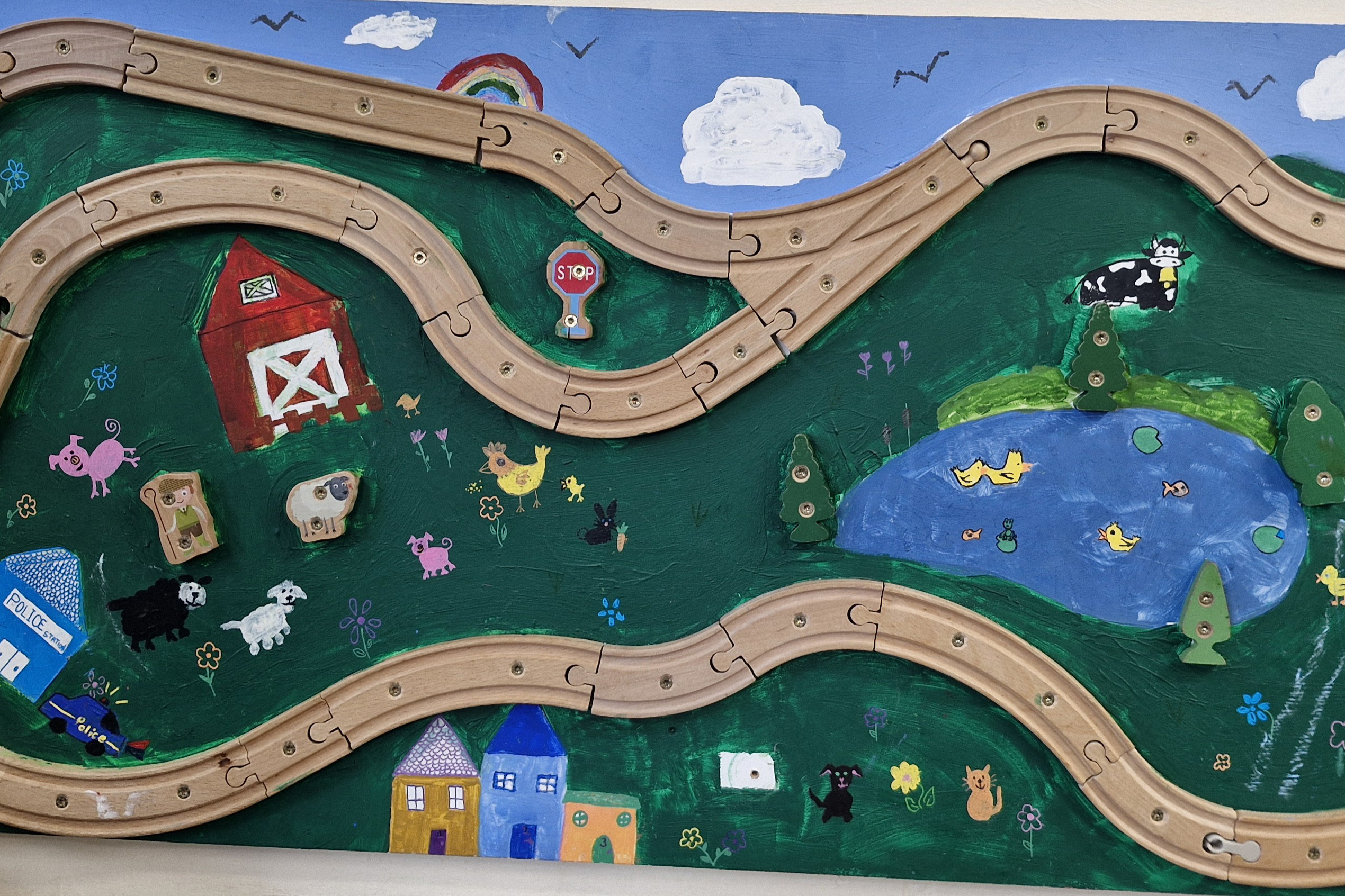 A board has been painted with fields, flowers, animals, a pond and houses. There is a wooden track going around the board.