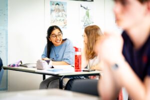 A student sits at a classroom table with another student. They are midway through conversation and they have pens, notebooks, pencilcases and water bottles in front of them.