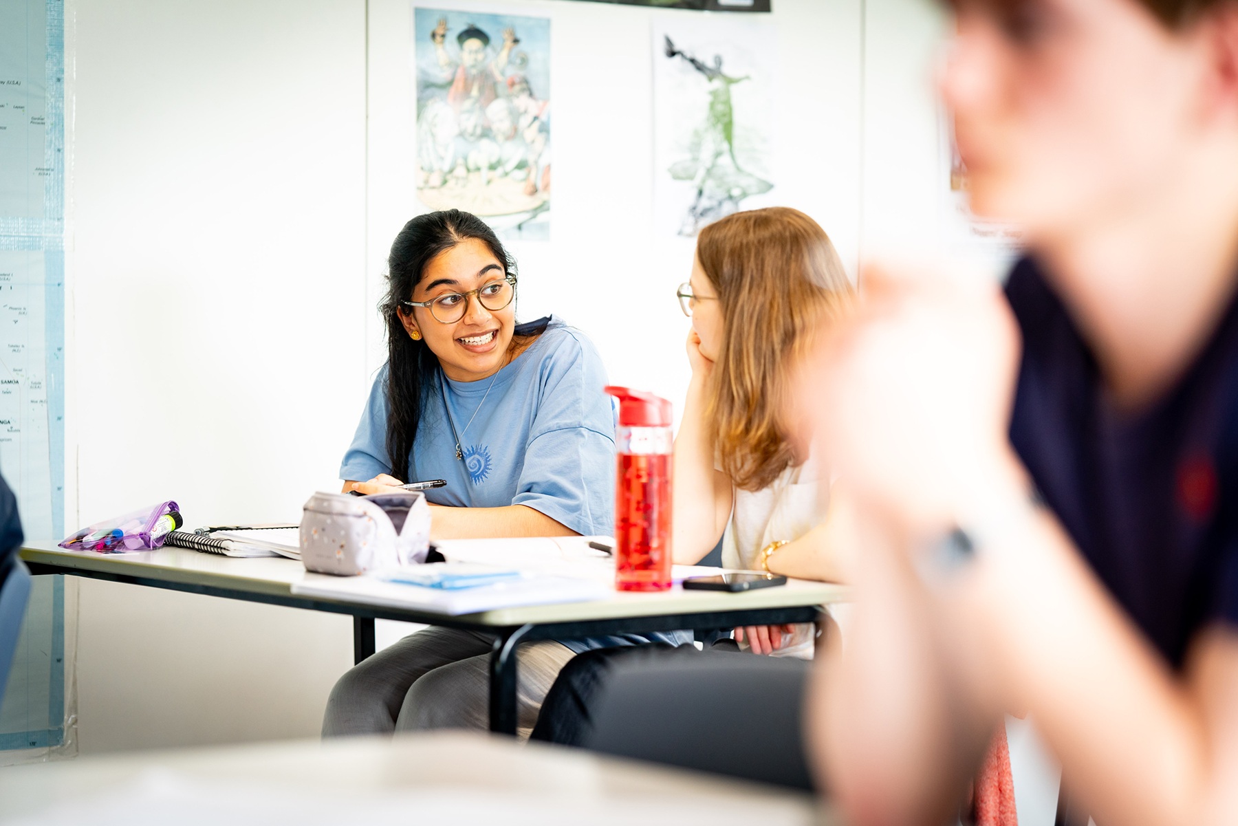A student sits at a classroom table with another student. They are midway through conversation and they have pens, notebooks, pencilcases and water bottles in front of them.