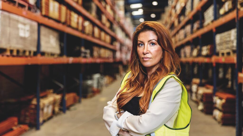 An image of a woman who is looking at the camera with her arms folded. She has red hair and is wearing a high vis jacket.