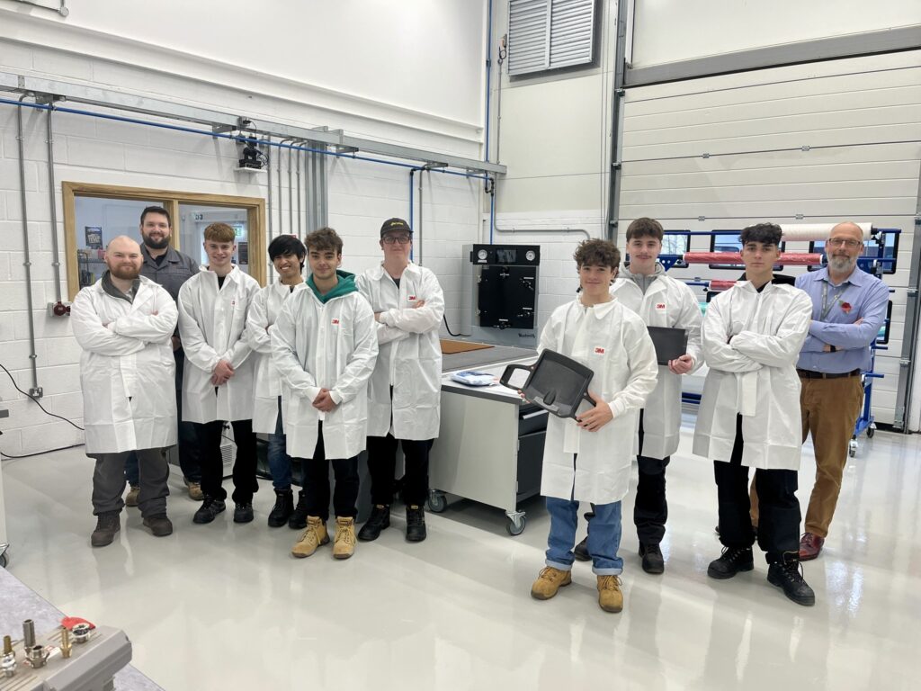 A group of students in lab coats face the camera in the engineering bay of the STEM & Health Skills Centre.