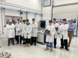 A group of students in lab coats face the camera in the engineering bay of the STEM & Health Skills Centre.