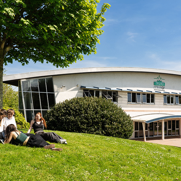 Students sat on the grass under a tree outside the Helford building on a blue sky day.