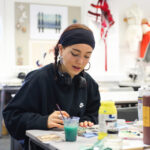 A girl in a black sweatshirt sits in an Art and Design classroom. She has a paintbrush in hand and has paints surrounding her on the table.