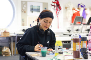 A girl in a black sweatshirt sits in an Art and Design classroom. She has a paintbrush in hand and has paints surrounding her on the table.