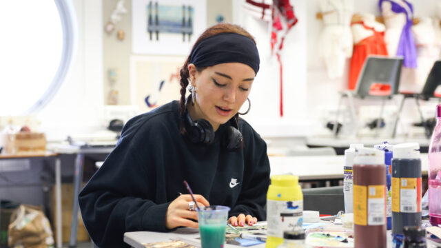 A girl in a black sweatshirt sits in an Art and Design classroom. She has a paintbrush in hand and has paints surrounding her on the table.