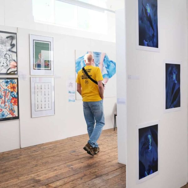 A person walks through an art exhibition. There are paintings and drawings up on plain white walls.