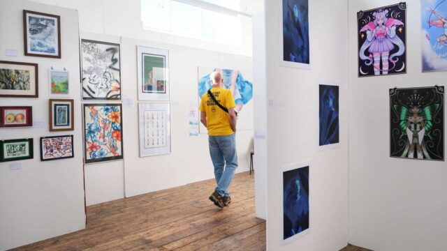 A person walks through an art exhibition. There are paintings and drawings up on plain white walls.
