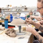 A close up of an adult male working with ceramics. He has clay on a table in front of him and he is using a small scalpel on the clay. The sleeves of his blue fleece are rolled up.