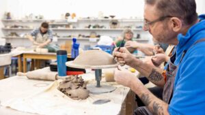 A close up of an adult male working with ceramics. He has clay on a table in front of him and he is using a small scalpel on the clay. The sleeves of his blue fleece are rolled up.