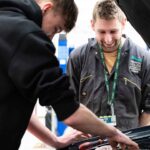 A tutor from the automotive department watches over a student as he works underneath the bonnet of a car.