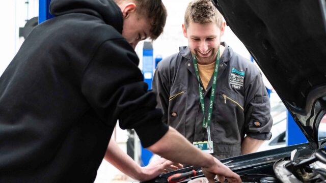 A tutor from the automotive department watches over a student as he works underneath the bonnet of a car.
